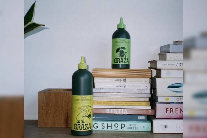 two bottles of Graza olive oil, one sitting next to books and the other on top of books