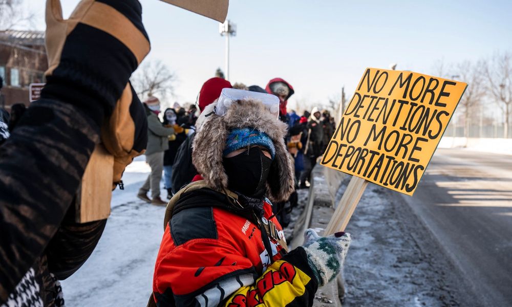 Masked protester holds sign reading "No More Detentions No More Deportations"