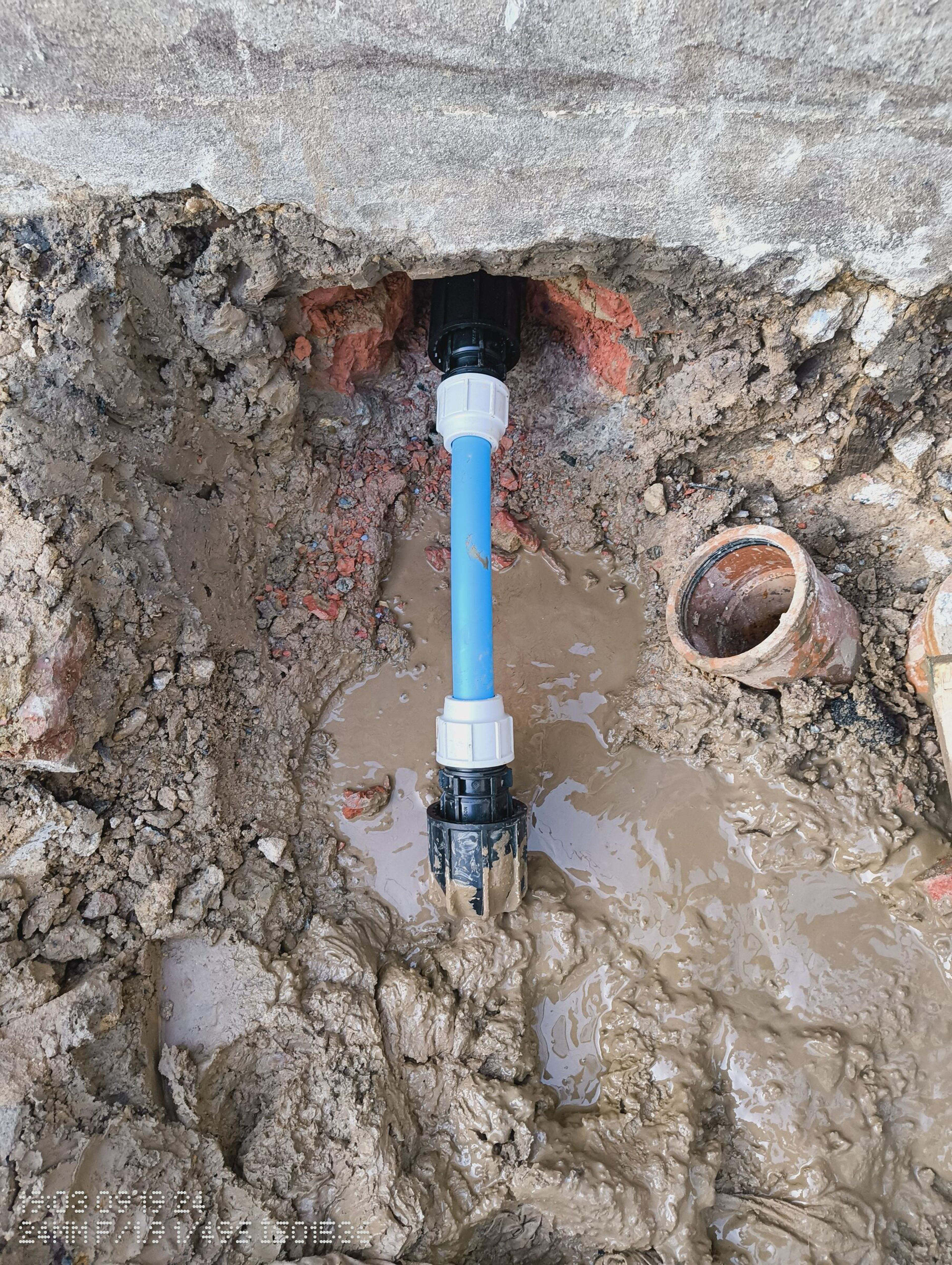 Existing water pipe with compression fittings standing in muddy ground under the concrete edge, beside a cut drain.