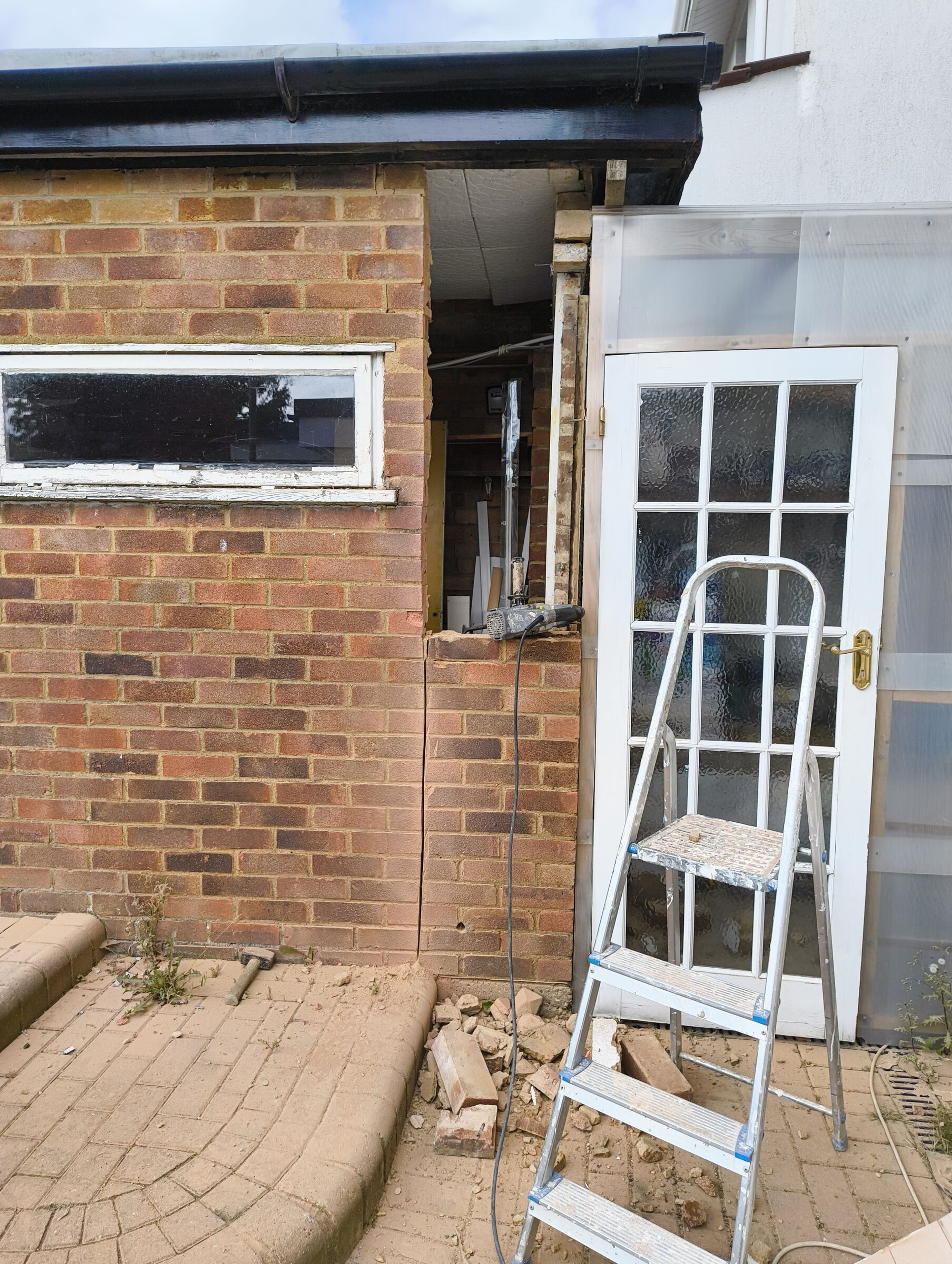 Opening cut through the brick wall of the rear extension, with rubble on the patio and a stepladder by the glazed door.