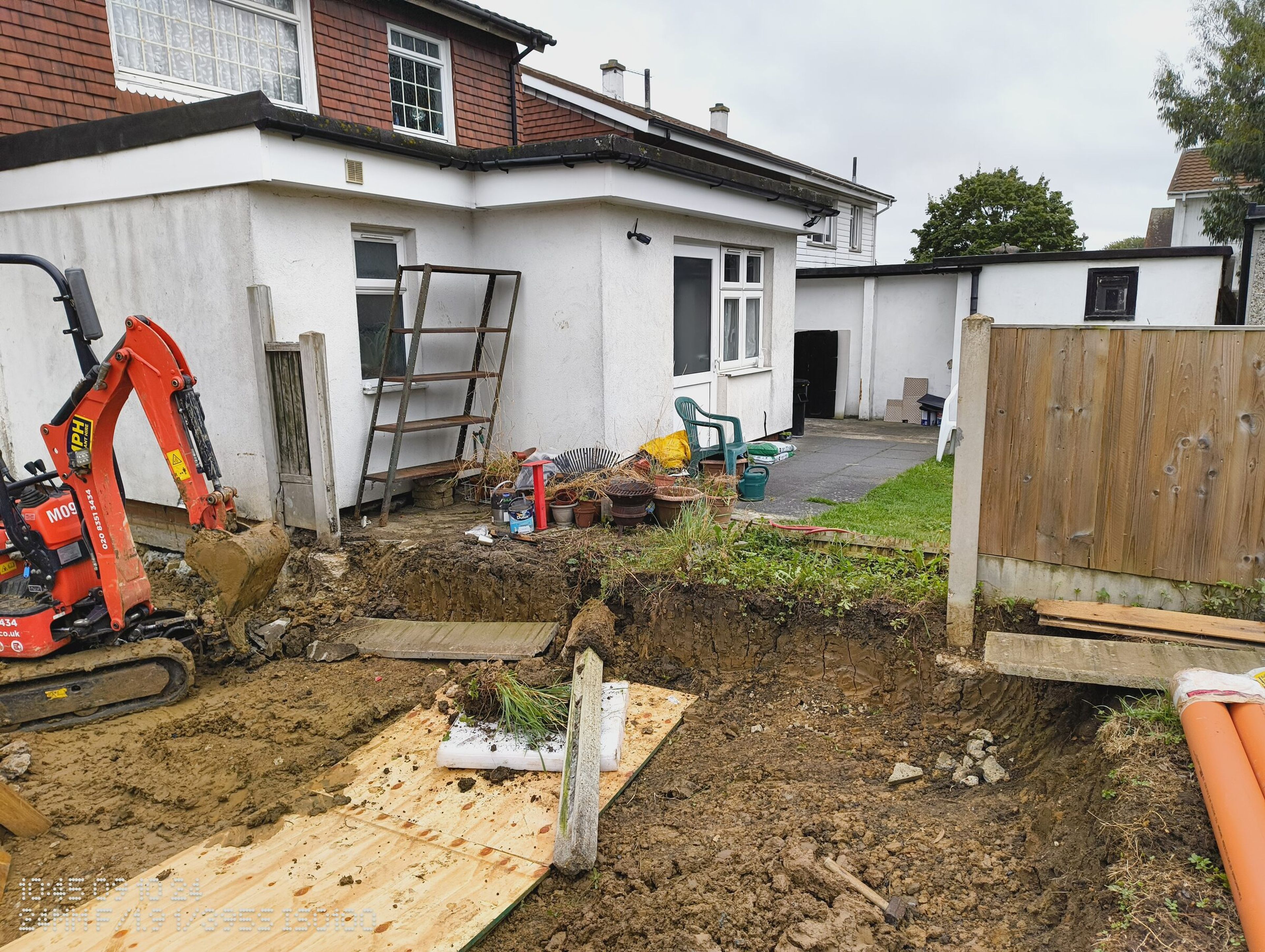 Mini digger starting the foundation dig along the side of a Chigwell house, with the garden edge cut back.