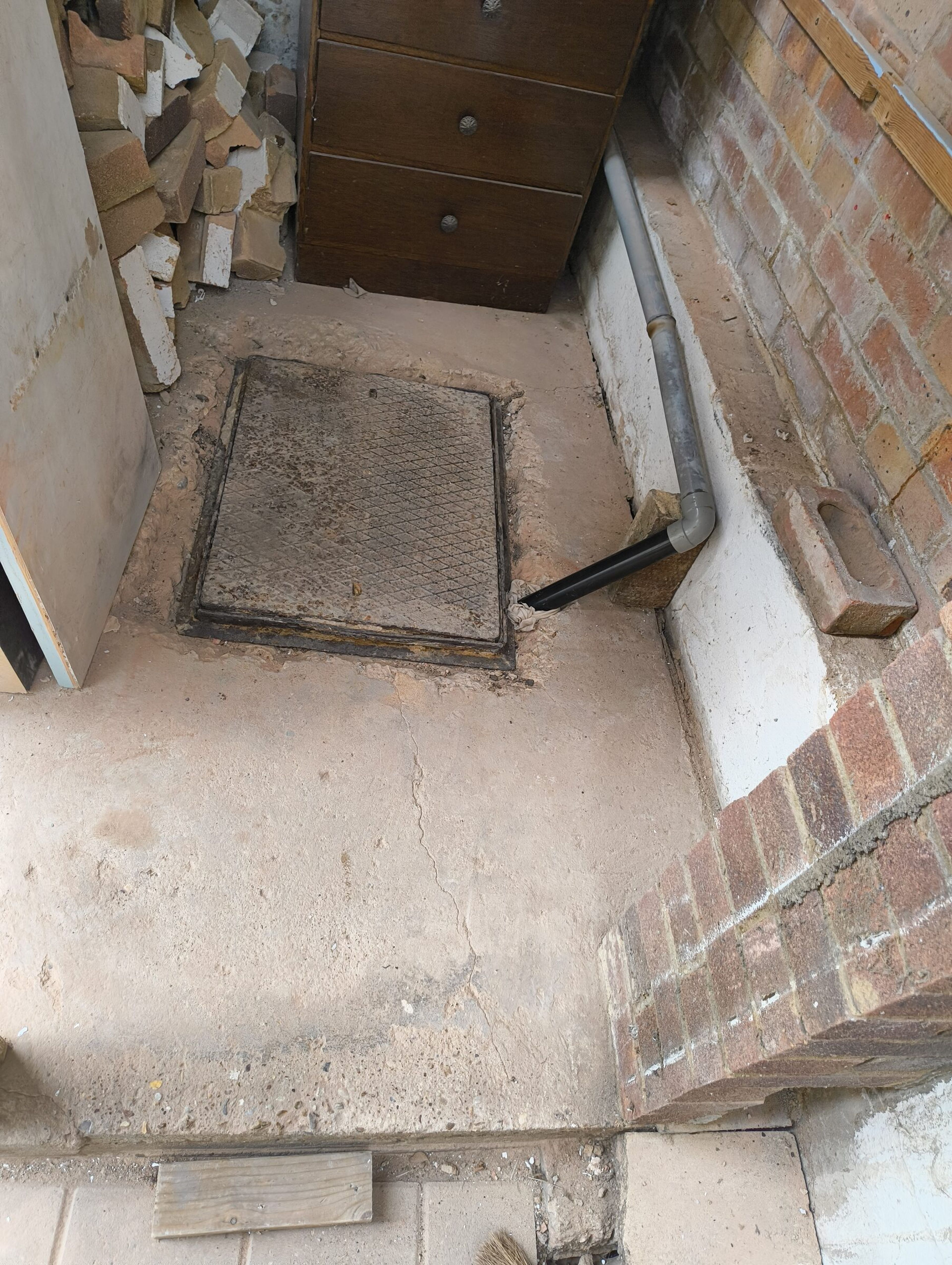 Metal manhole cover in the concrete floor beside a grey waste pipe and the external brick wall.