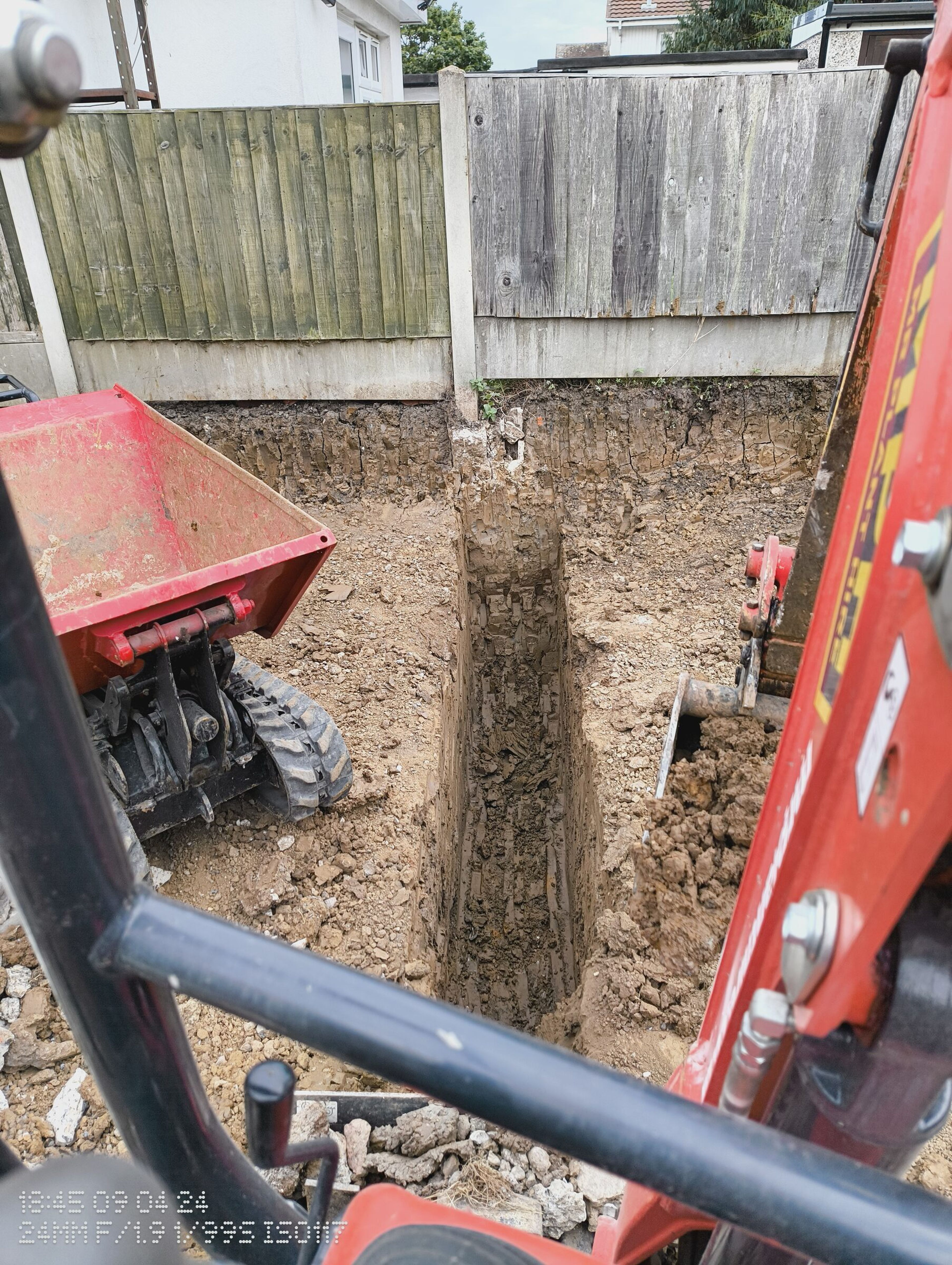Deep foundation trench dug beside a fence for a rear extension in Chigwell, with a mini digger and tracked dumper.