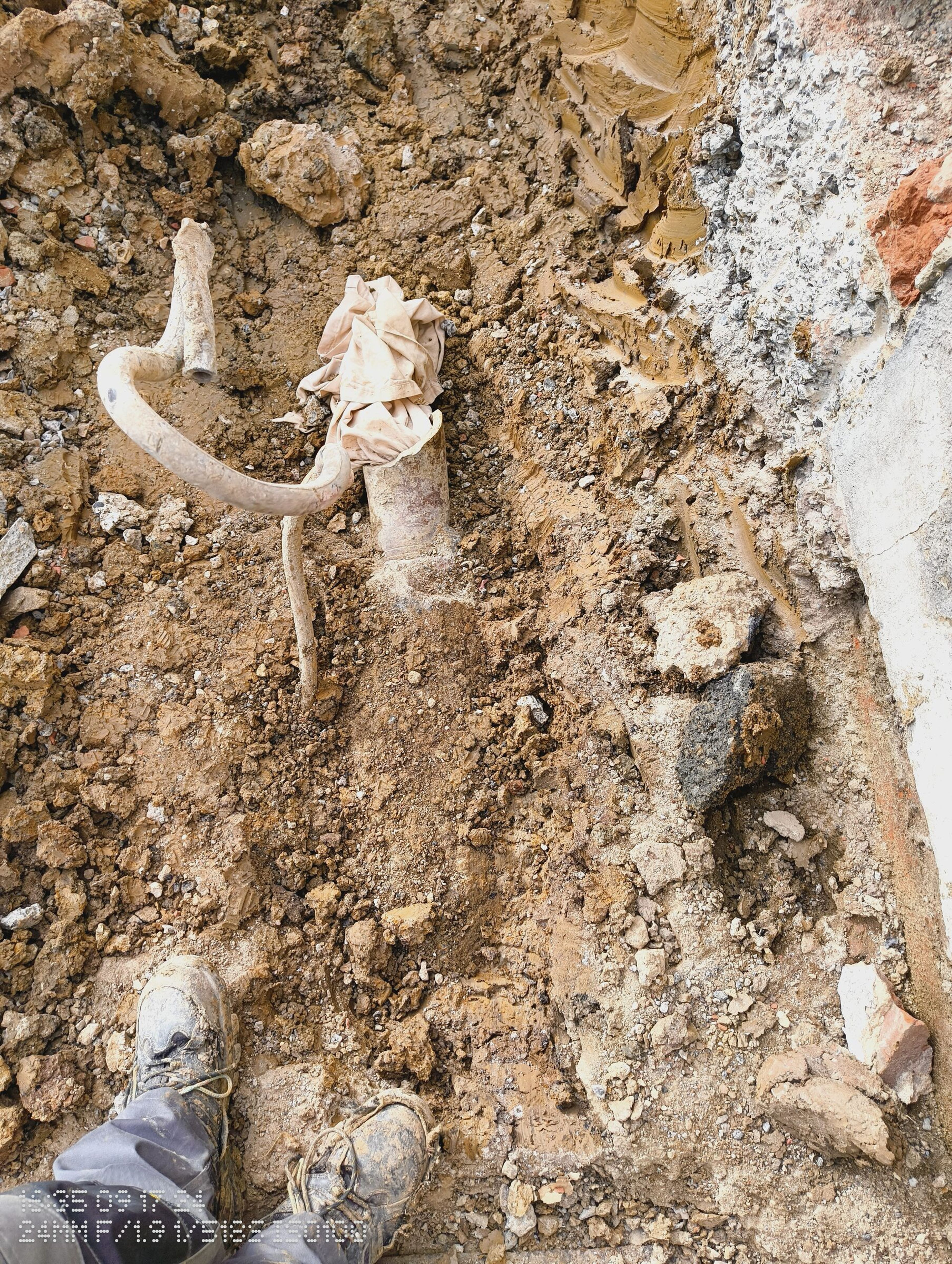 Curved pipe and a wrapped stub exposed in the clay next to the house wall.