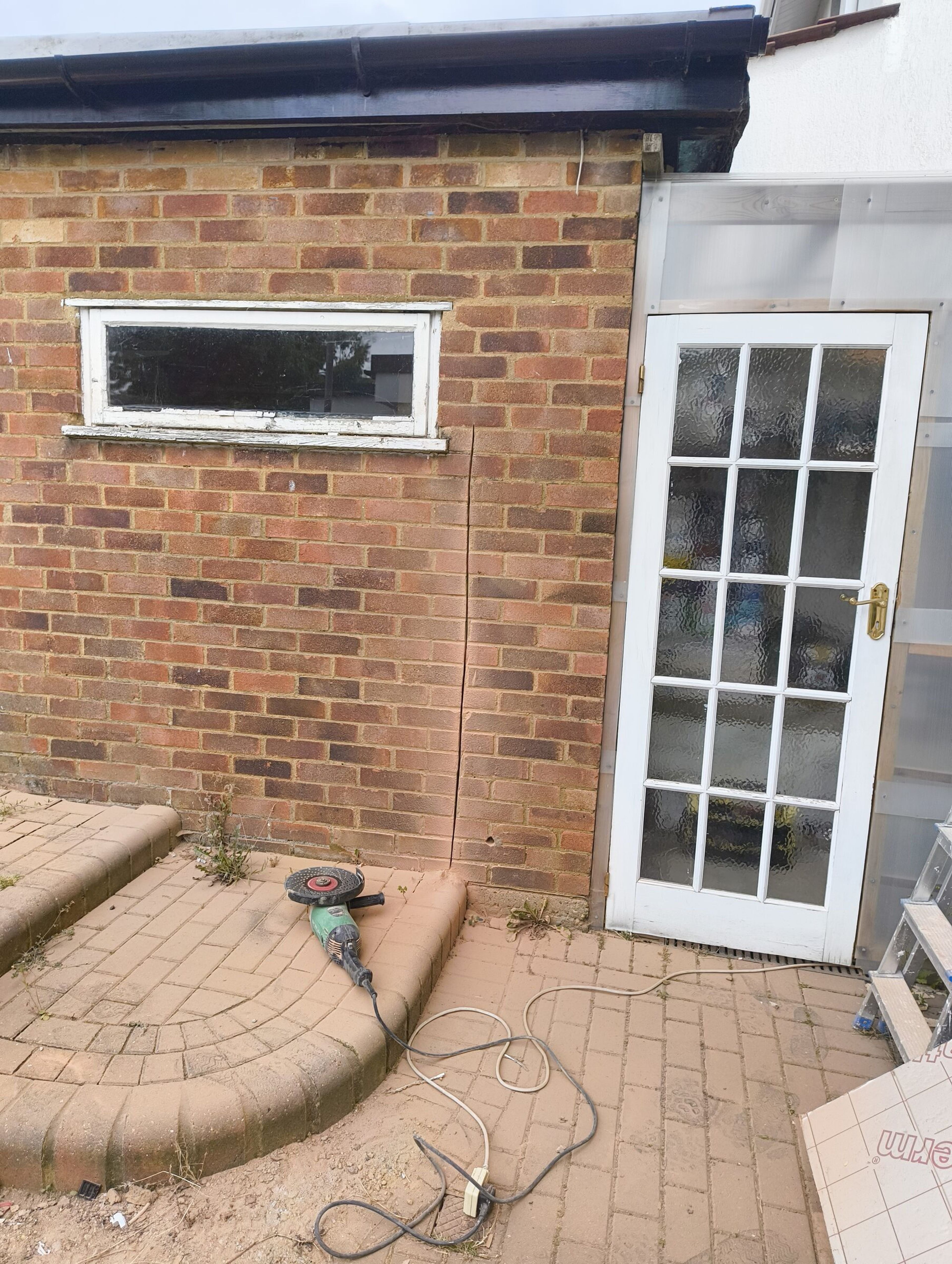 Brick rear extension in Chigwell before demolition, with a narrow window, a glazed side door, and an angle grinder on the patio.