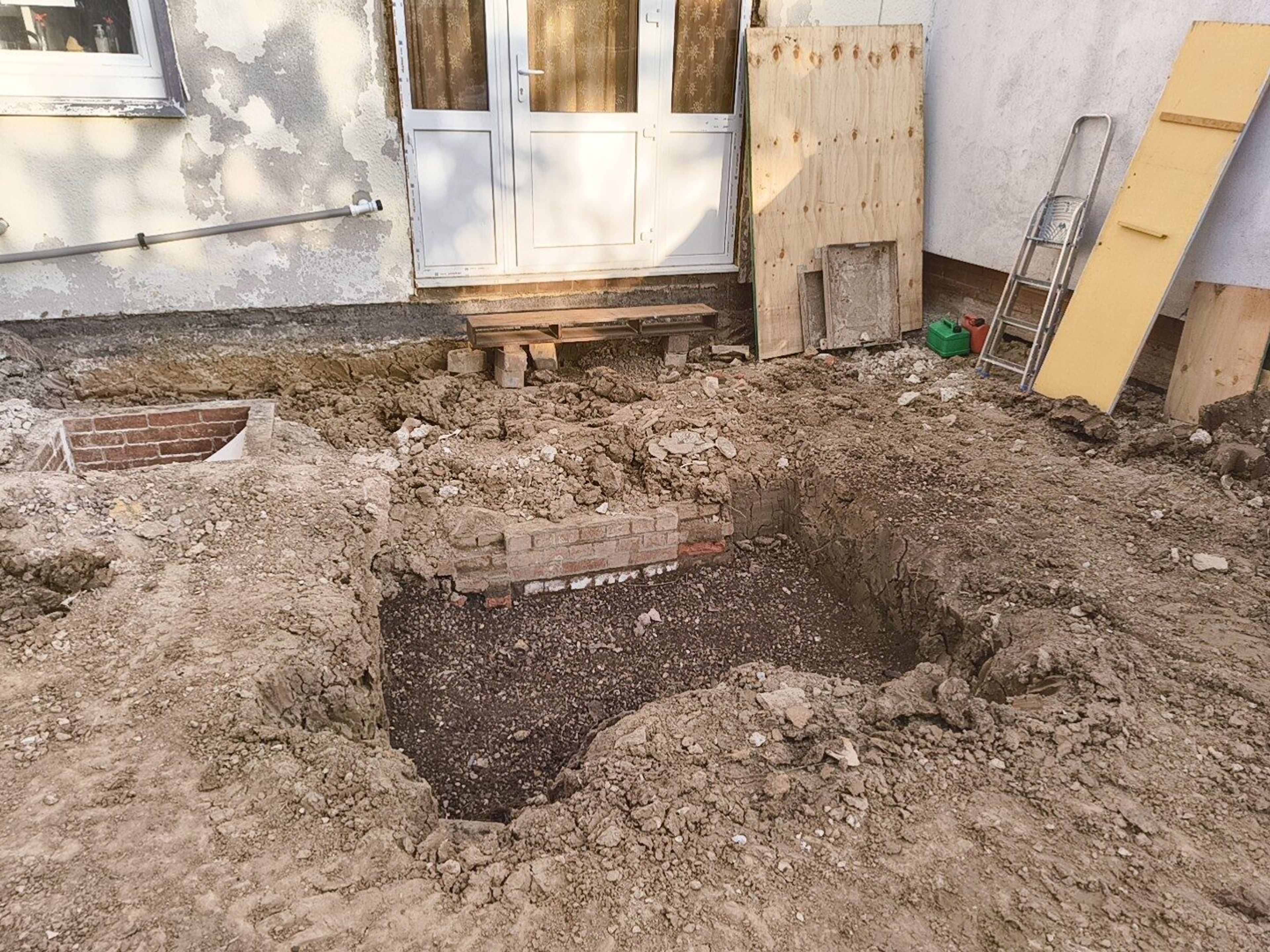 Excavated pit in front of the patio doors, with a low brick chamber and gravel in the bottom.