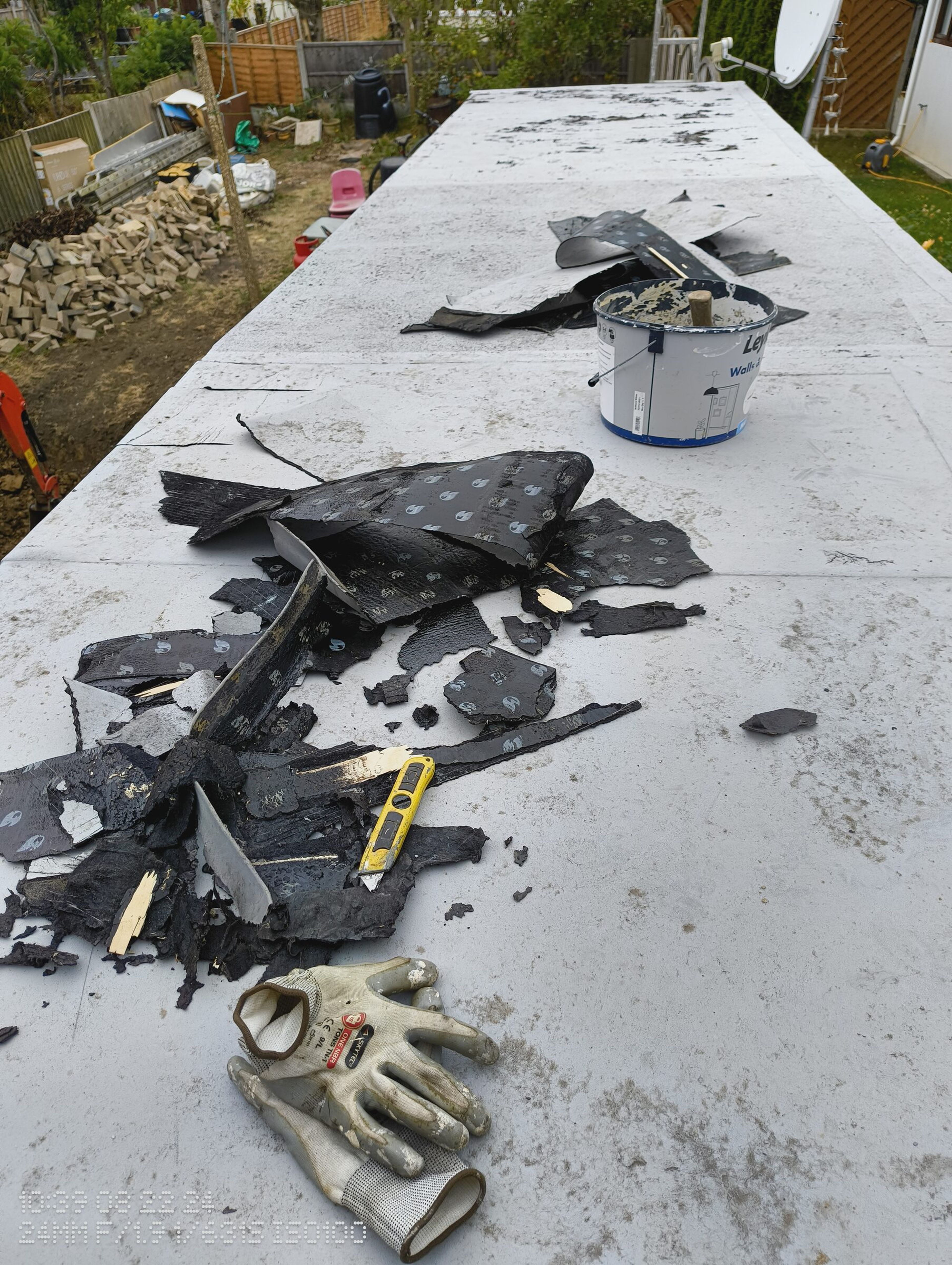 Flat roof with torn felt, a trimming knife, and work gloves, looking along the roof towards the garden.