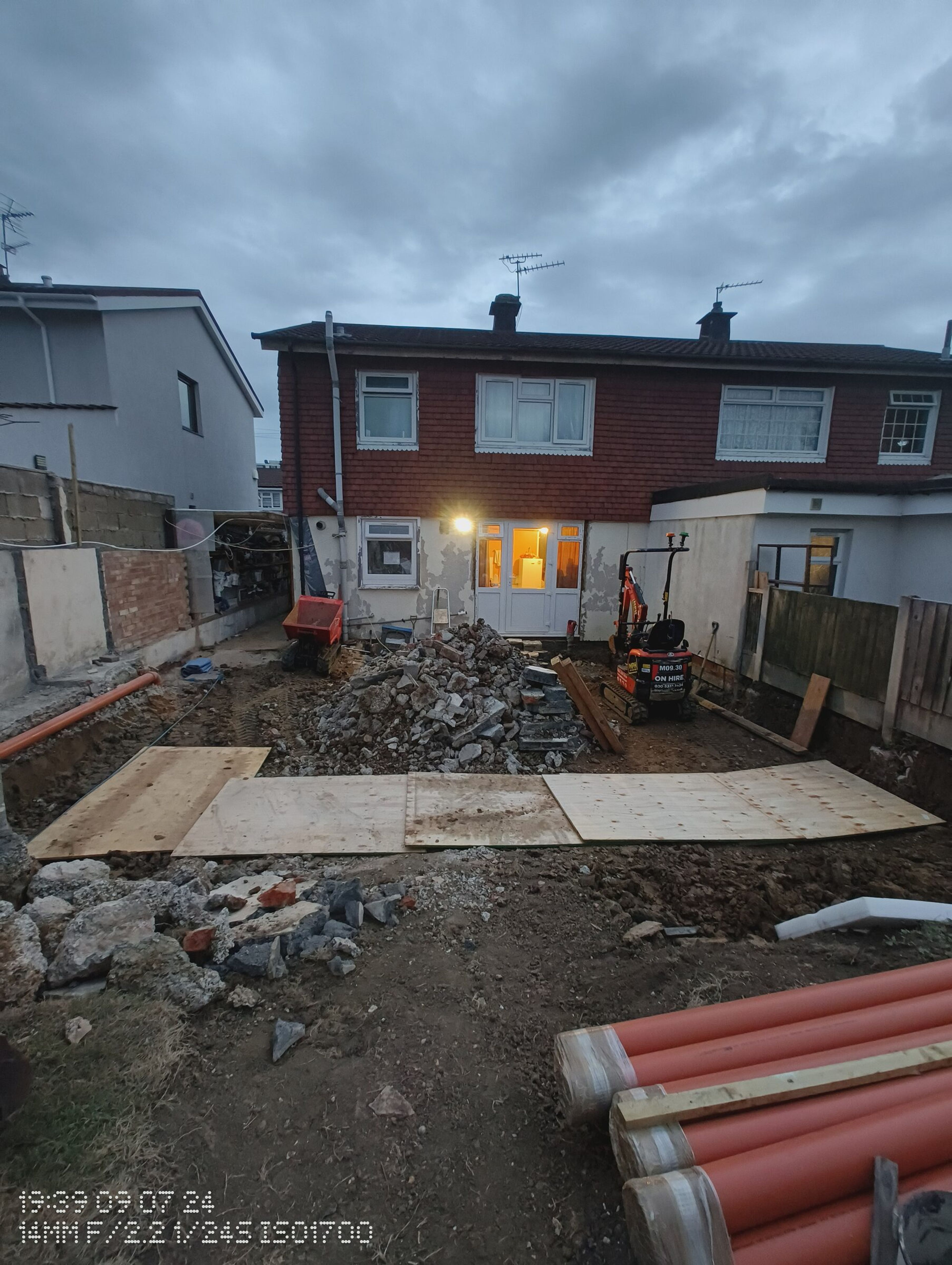 Evening view of a Chigwell rear extension site, with a mini digger, spoil heap and drainage pipes ready for the foundations.