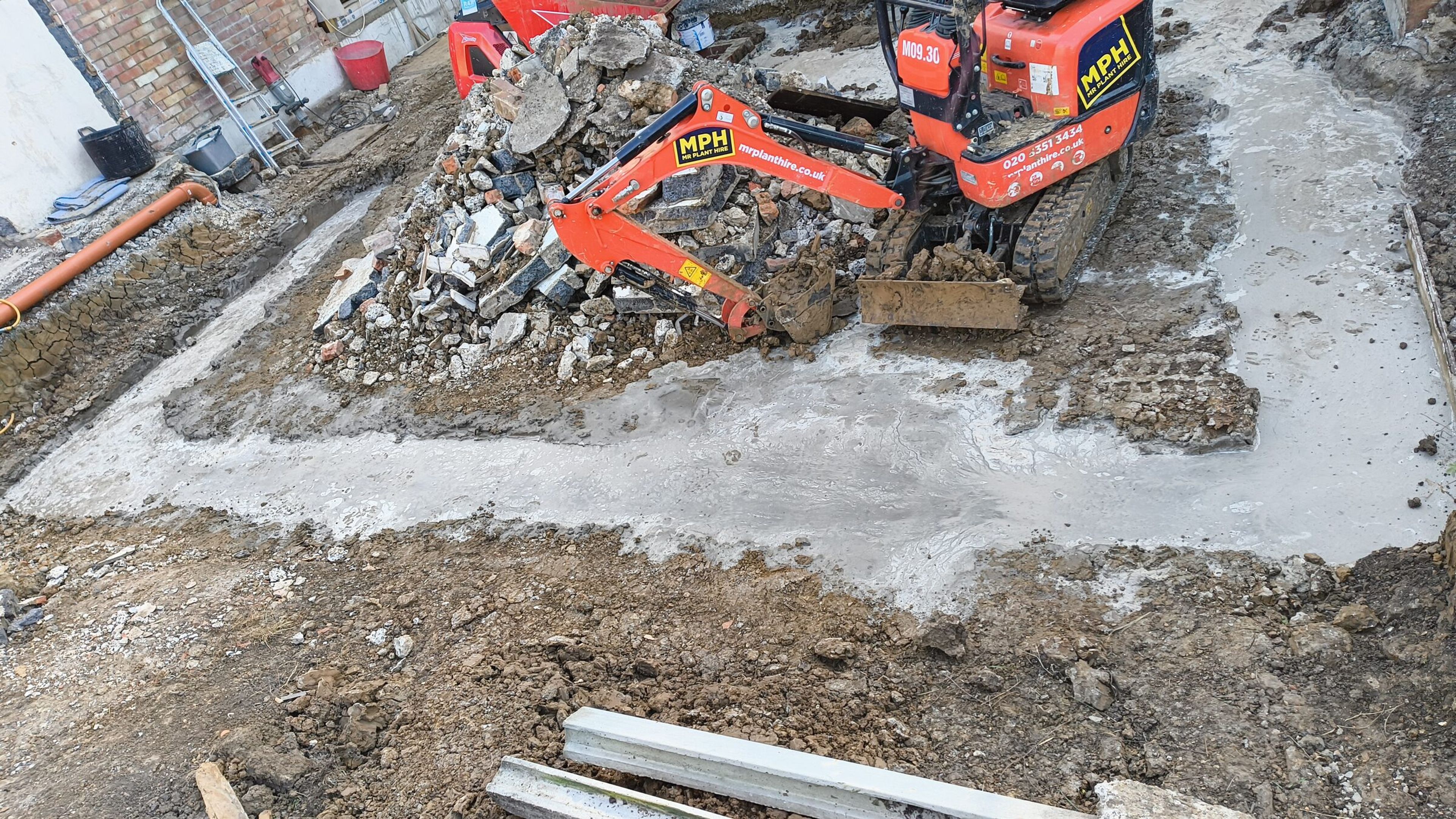 Mini digger beside a freshly concreted foundation trench and spoil heap on a Chigwell rear extension.
