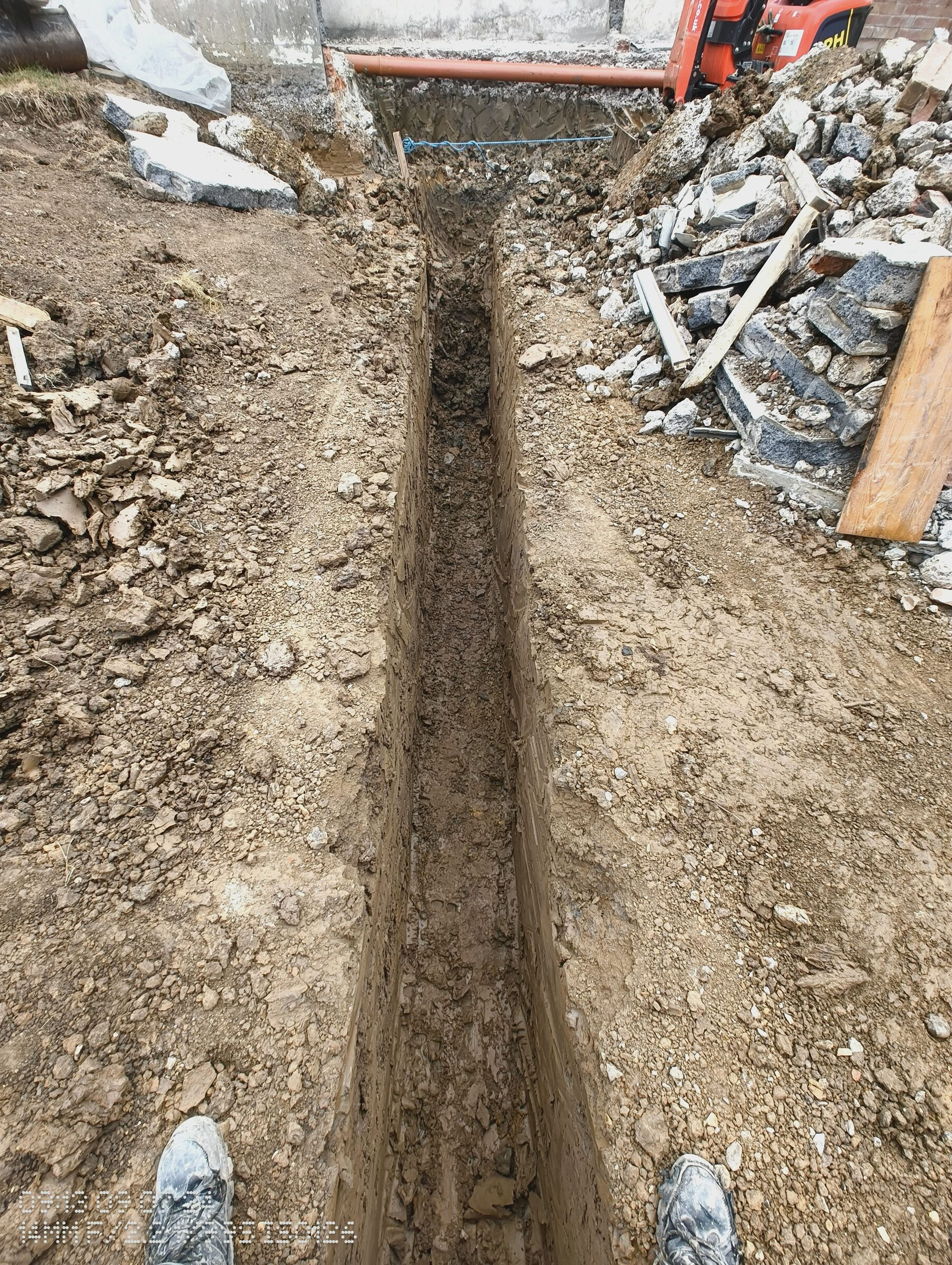 Foundation trench beside the house in Chigwell, with a drainage pipe in the excavation and spoil stacked to one side.