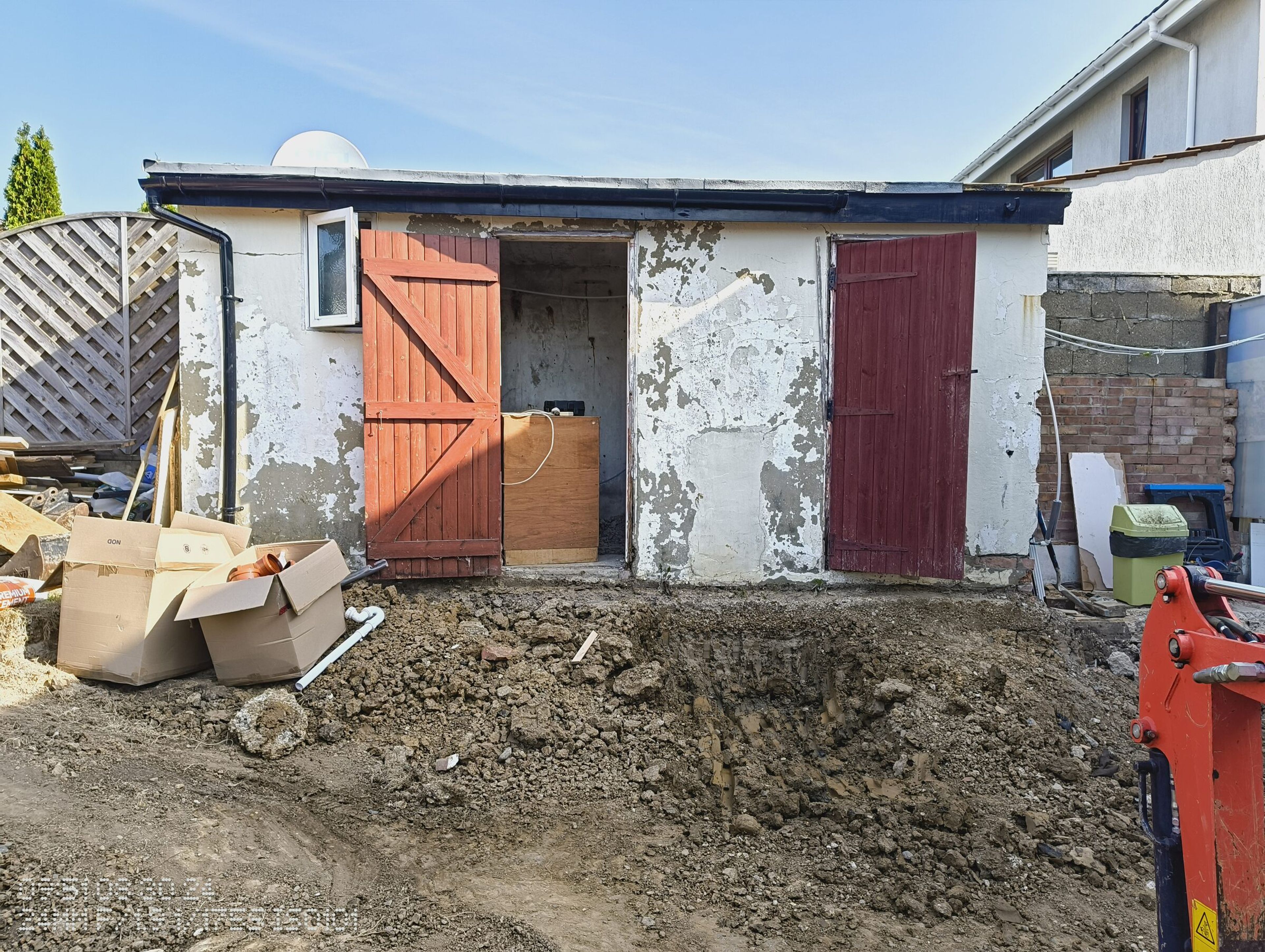 Rendered outbuilding with red doors at the back of the garden, with the ground dug out in front and a digger on site.
