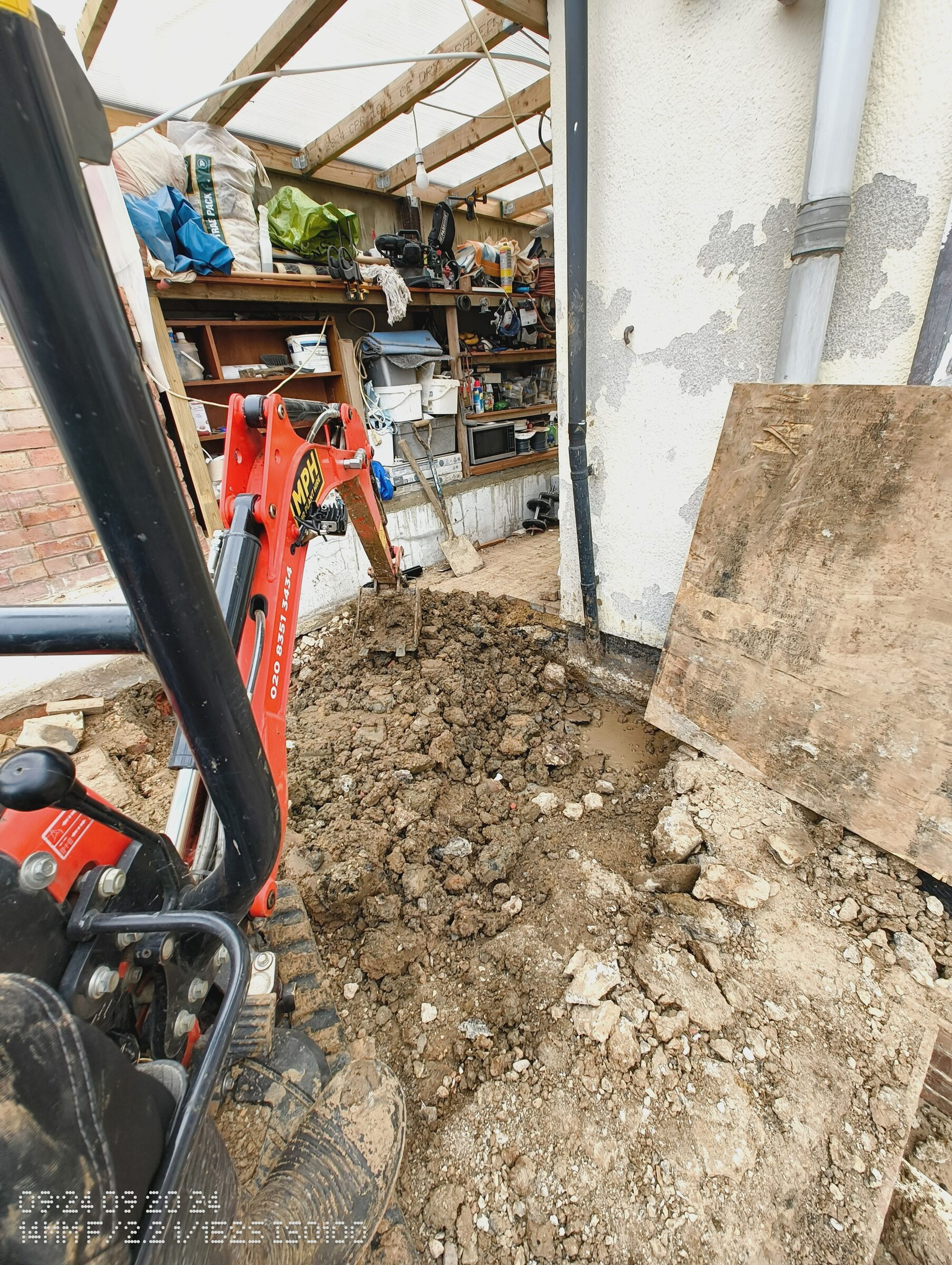 Mini digger at the muddy trench beside the house, with the soil pipe and a boarded-up opening.