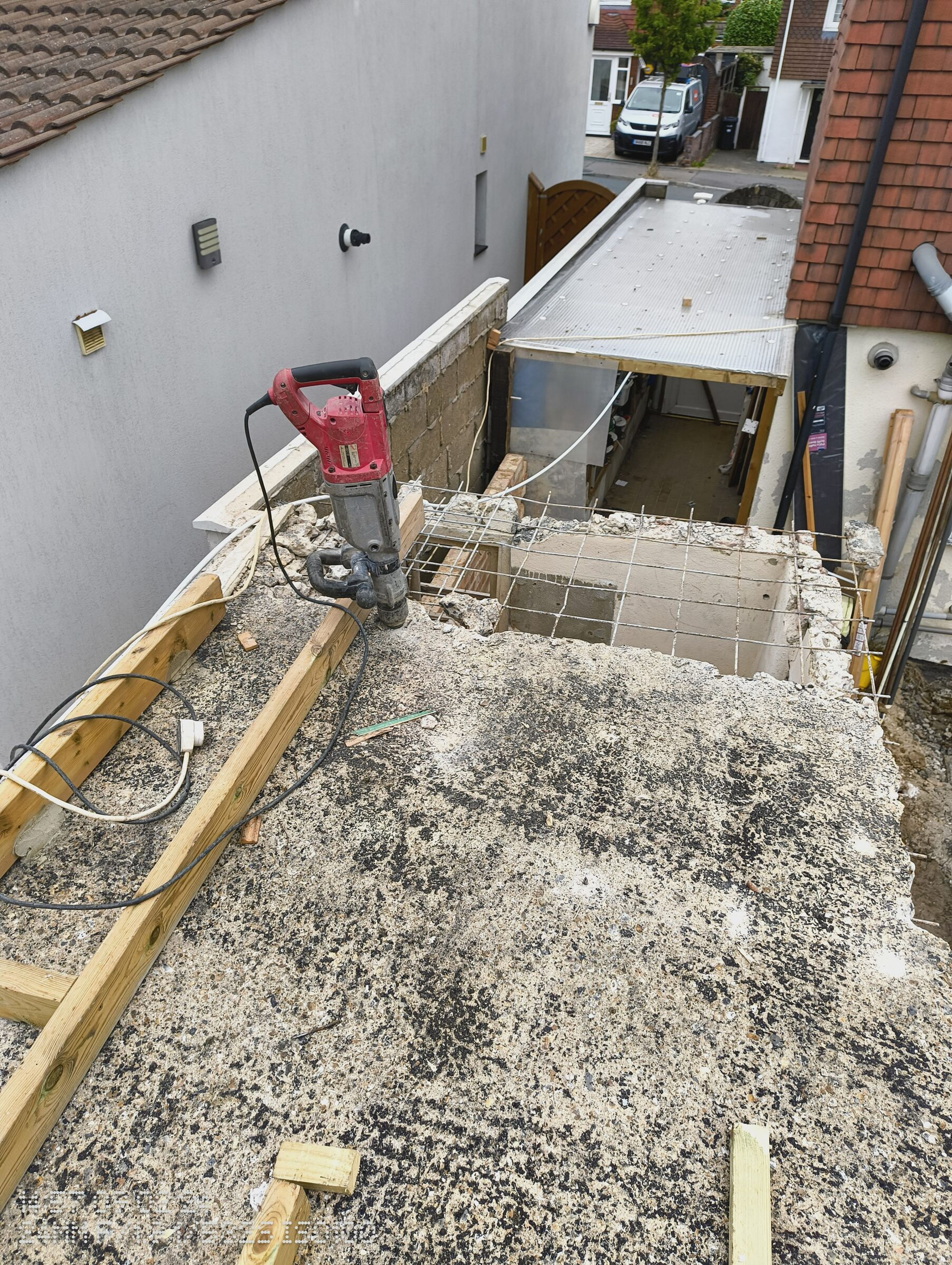 Breaker standing on the flat roof as the slab is broken out, with the side-return roof still in place beyond.
