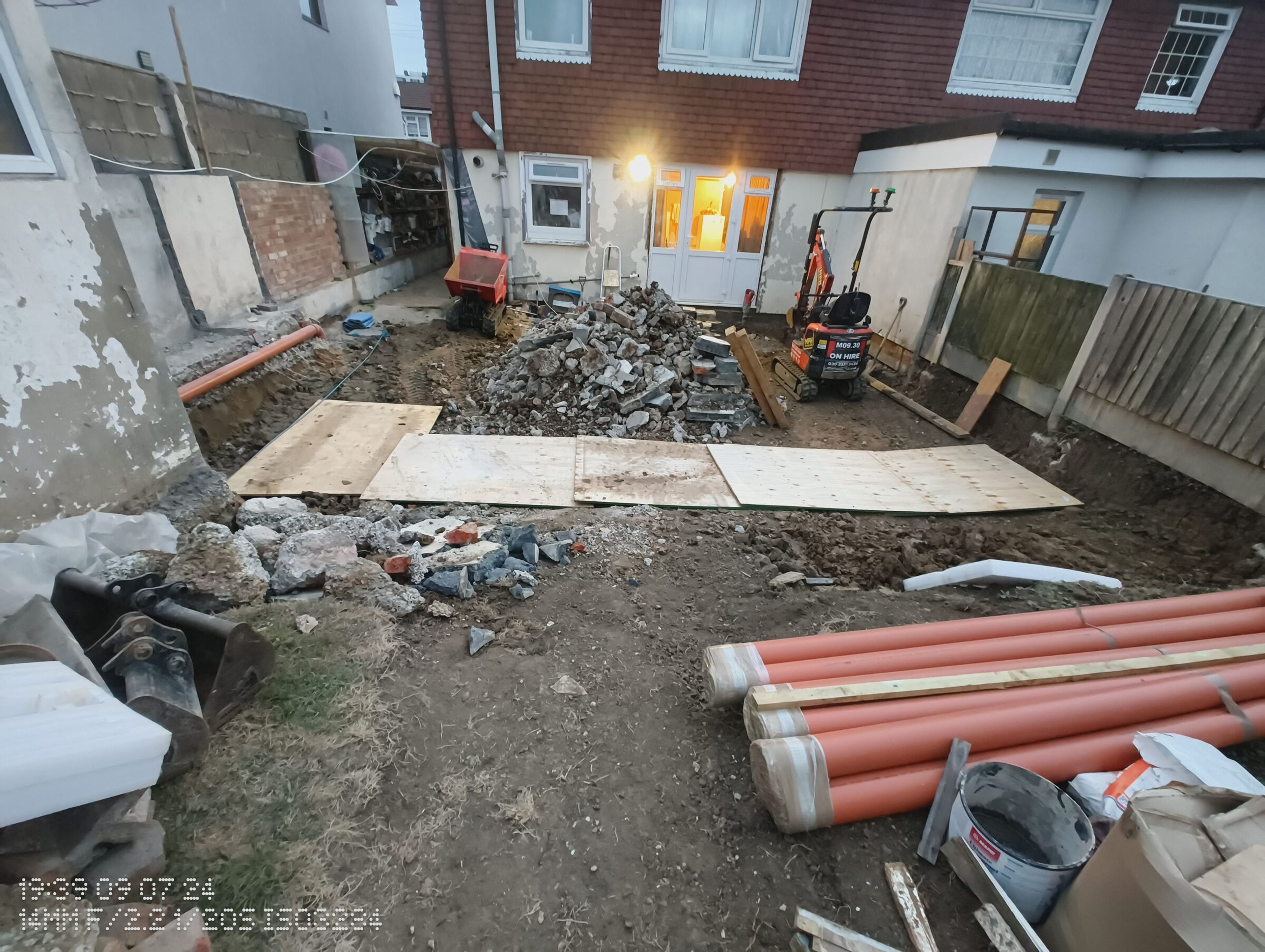 Rear garden in Chigwell set out for foundation work, with plywood boards, drainage pipes, a mini digger and a spoil heap.