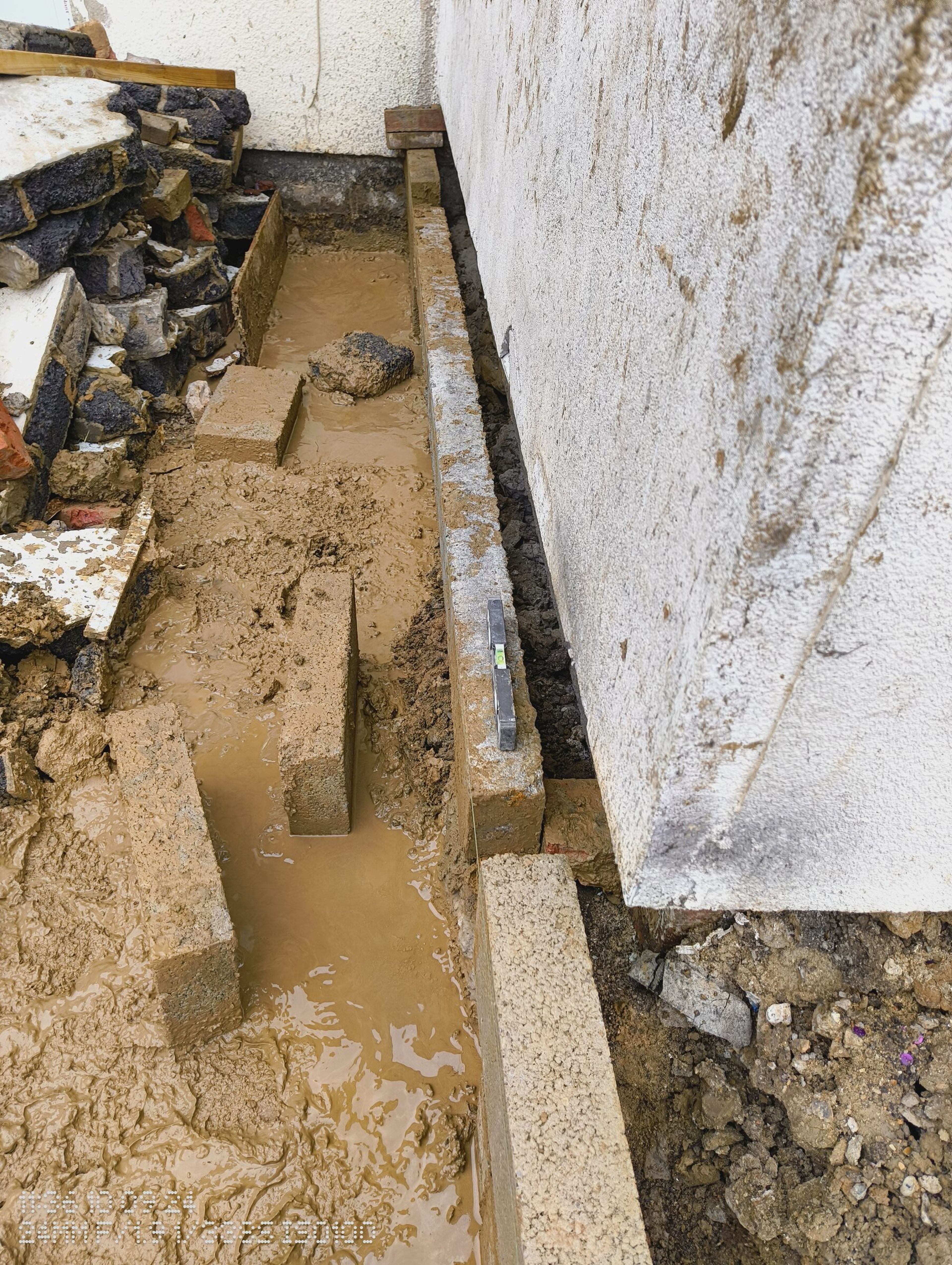 First courses of blockwork in the muddy trench beside the existing house wall.