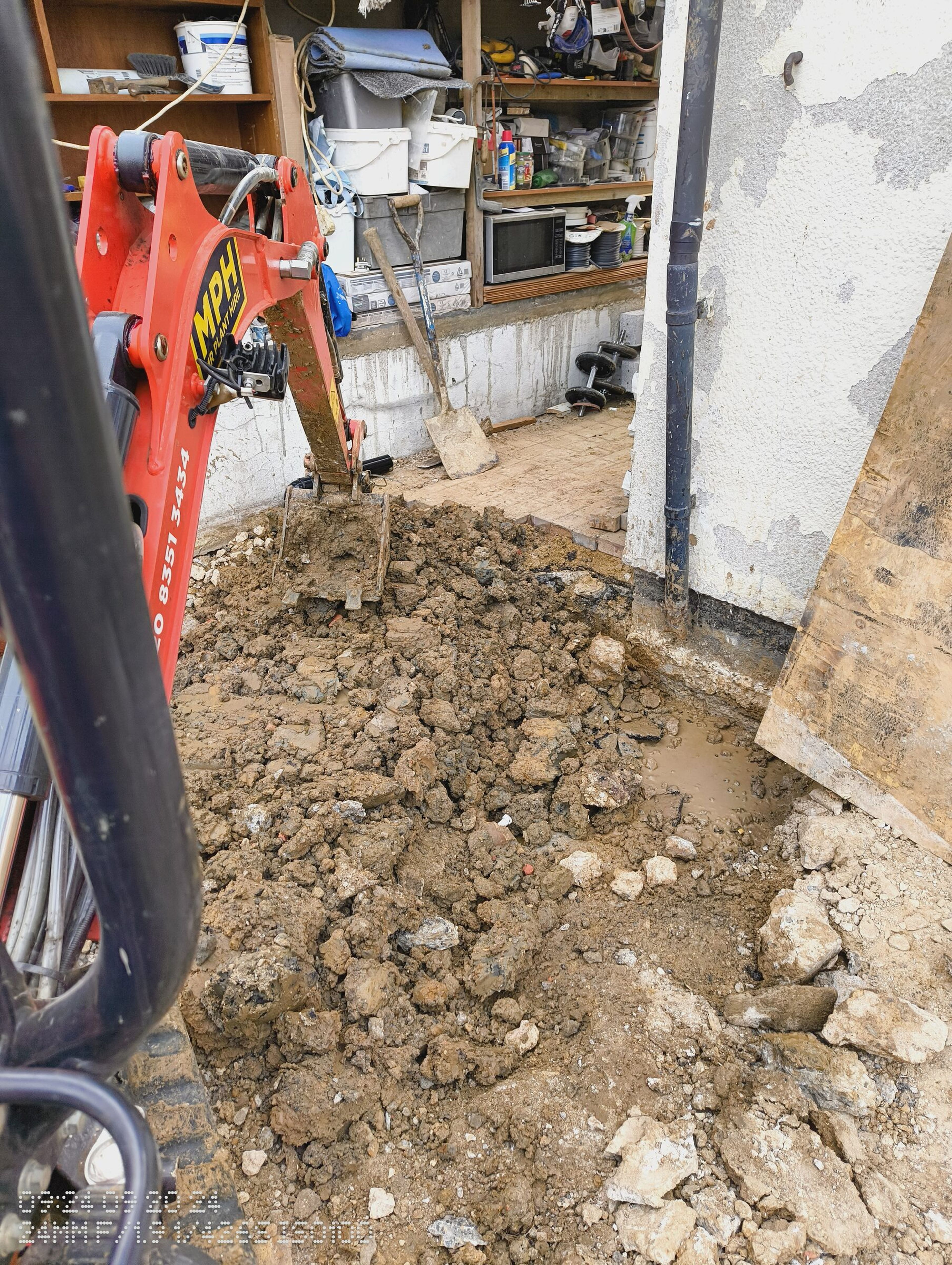 Mini digger beside the opened trench along the side of the house, with the workshop behind.