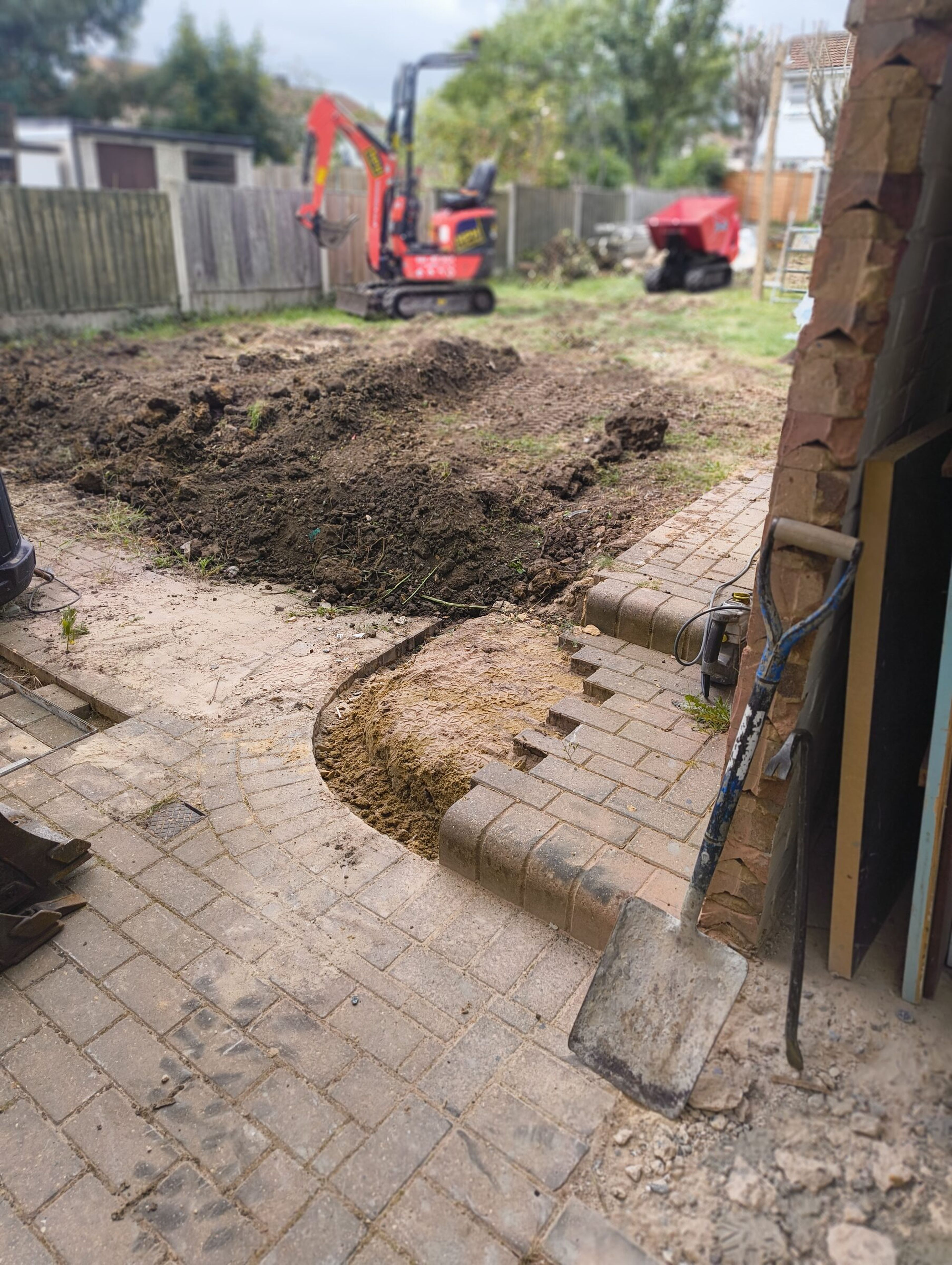 Patio partly lifted at the rear of the house, with a spoil heap, mini digger, and tracked dumper in the garden.