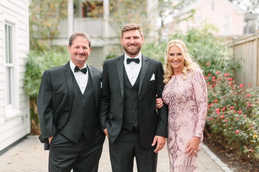 Two men in tuxedos and a woman in a pink beaded gown posing outside a historic Charleston SC home surrounded by roses