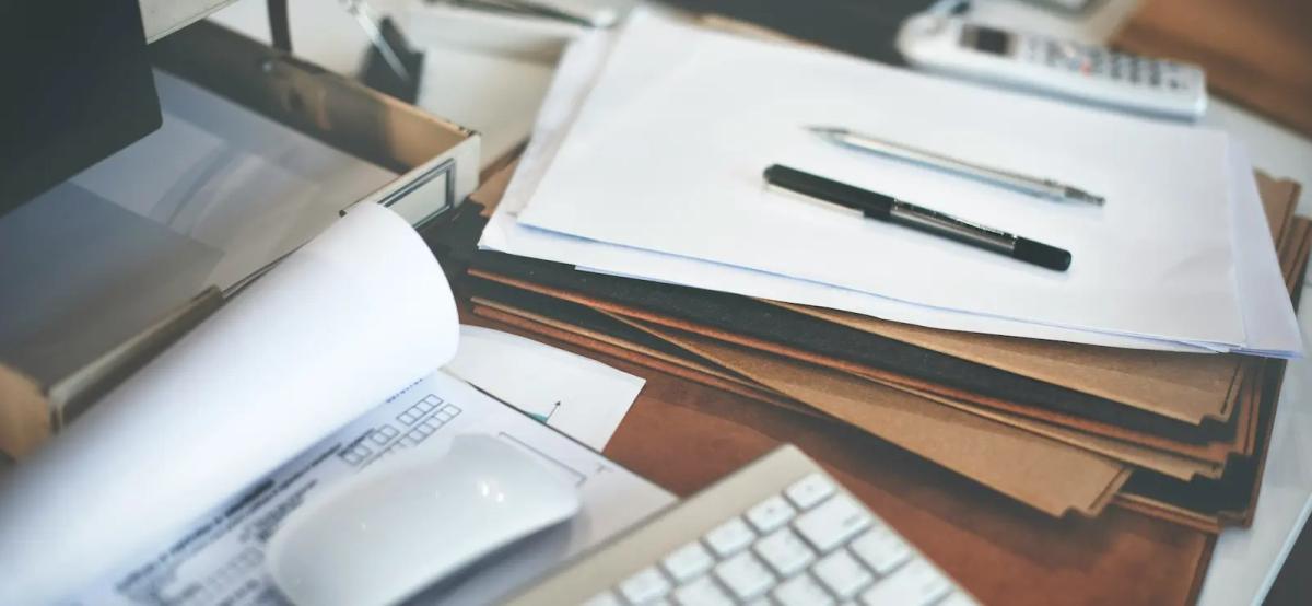 Cluttered office desk with piles of unsorted business documents and paperwork
