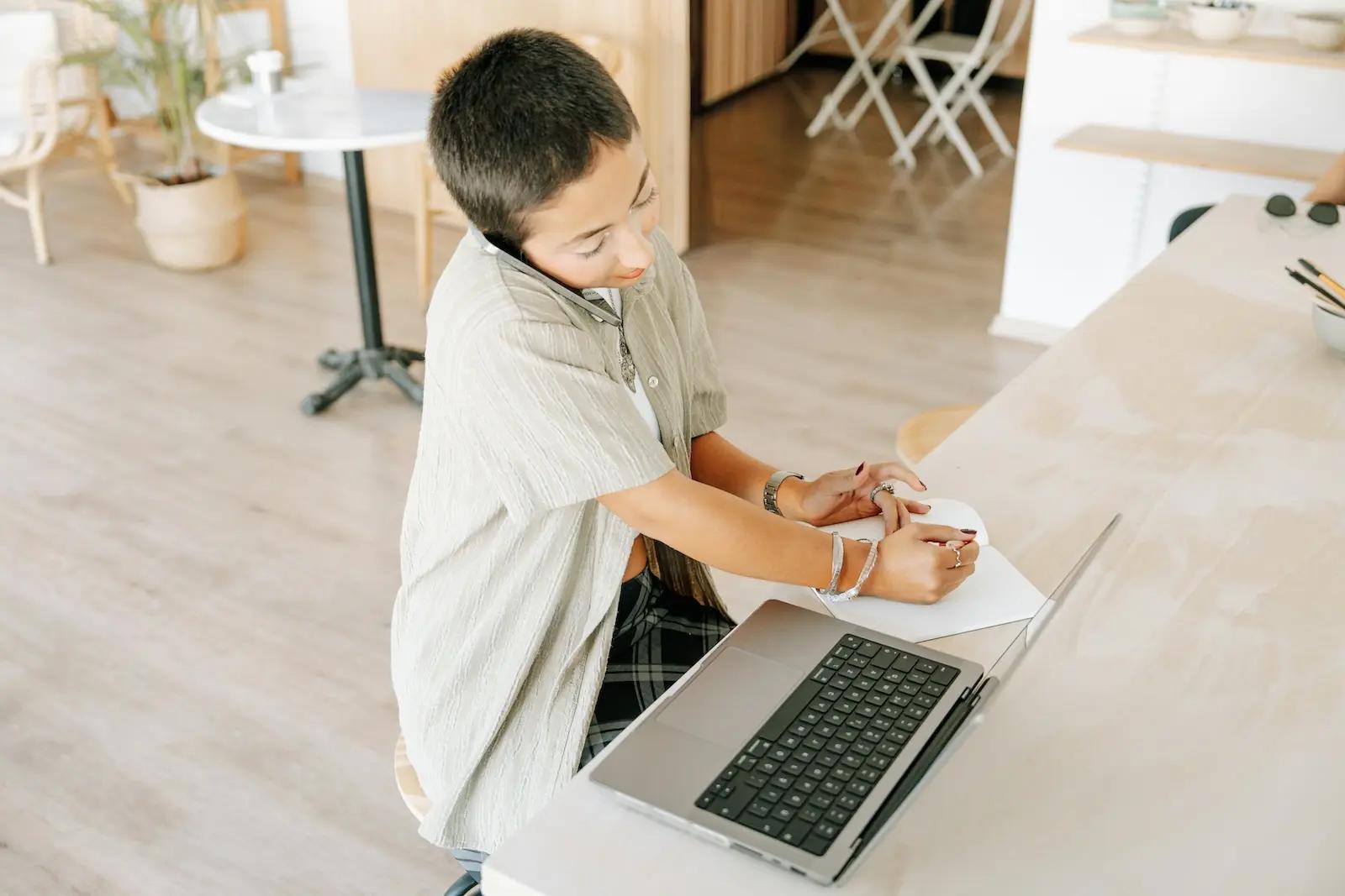 A professional talks on the phone while writing notes at a desk with a laptop, illustrating multitasking and work interruption in office environments.