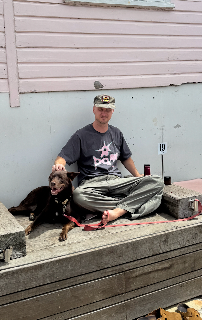 Founder bio picture of Sam Judd sitting barefoot and cross-legged on a wooden bench outdoors. He is casually petting an Australian red kelpie dog lying next to him on a red leash. Sam is wearing a cap, a dark graphic t-shirt, and loose green pants, and is squinting at the camera—either from the bright sun or because he doesn't like having his photo taken - more likely the latter. Behind them is a building with white panels and pink weatherboard siding, a fresh beetroot juice and a table marker with the number 19 sits nearby.