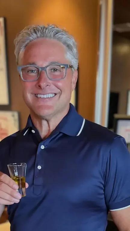 Ralph Hersom smiling in glasses and a navy polo shirt, holding a small glass indoors.