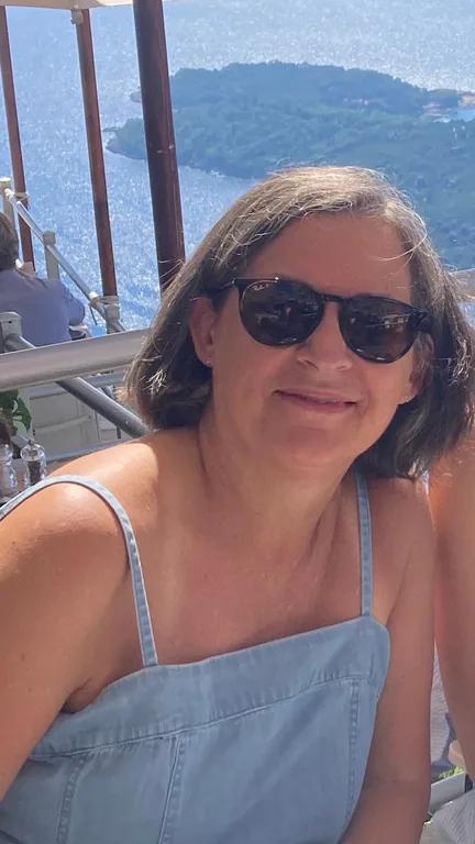Kim Trowbridge wearing sunglasses and a pale blue top on a terrace overlooking the sea.