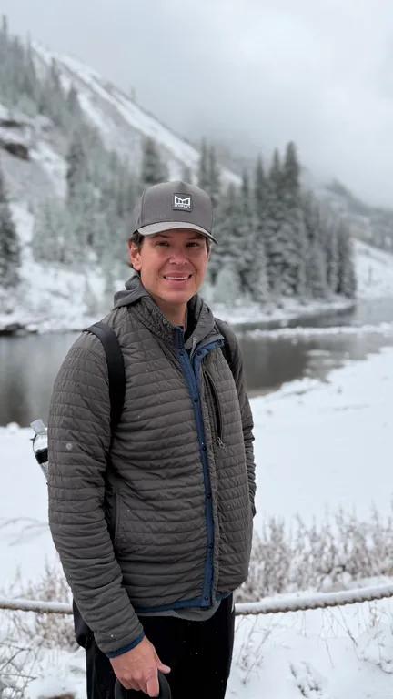 Travis Chick smiling in a cap and dark vest beside a snowy lake and forest.