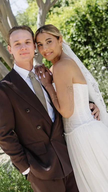 Lauren Noelle in a wedding dress beside her partner beneath leafy trees.