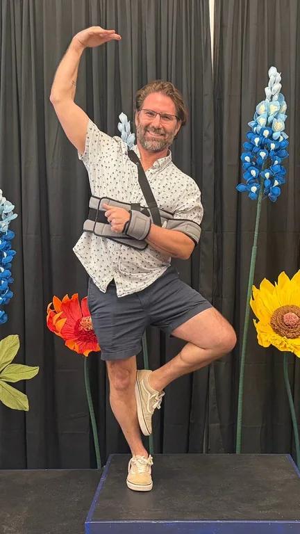Mason Arnold smiling and balancing on one leg with an arm in a sling, in front of colorful paper flowers.