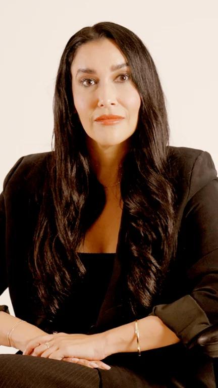 Rebecca seated in a black outfit, with long dark hair against a light background.