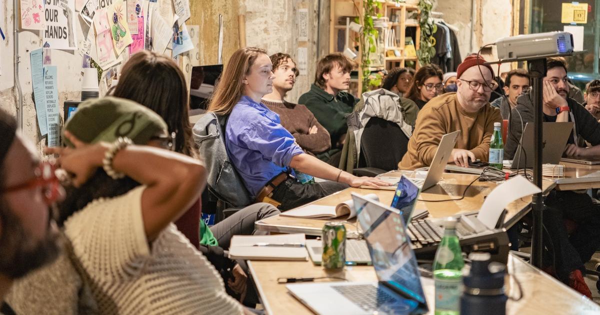 A group of people seated at long tables with open laptops, attending a workshop or presentation.
