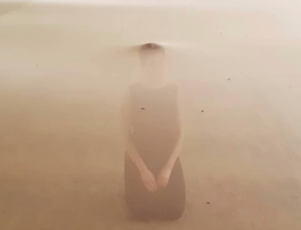 A kneeling figure in a dark dress, almost submerged in a light brown, hazy medium.