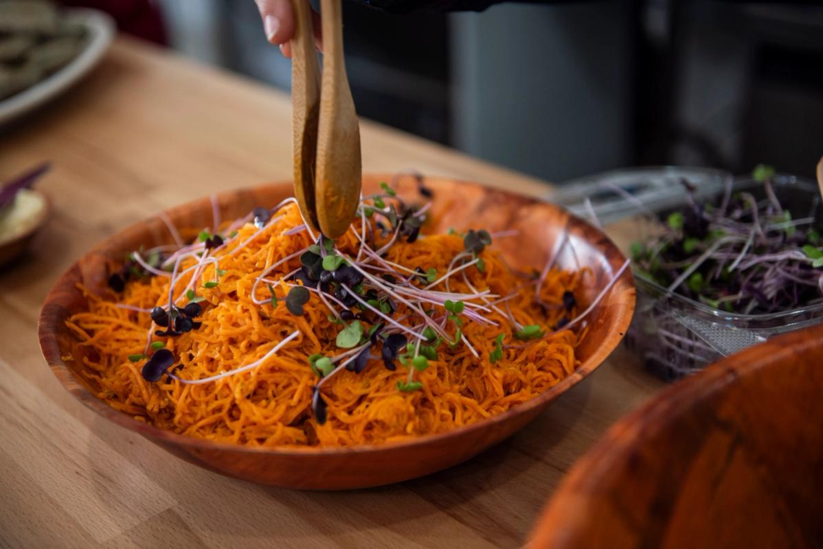 A hand with wooden tongs hovers over a wooden bowl of shredded carrots and microgreens.