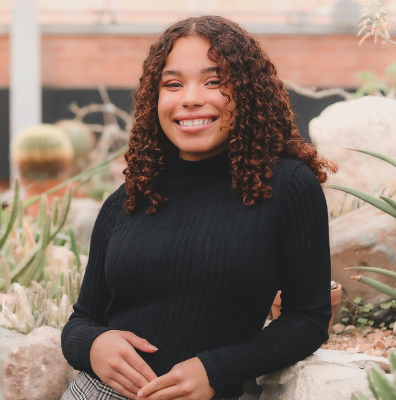 Smiling young woman with curly hair in a black turtleneck, surrounded by cacti and succulents.