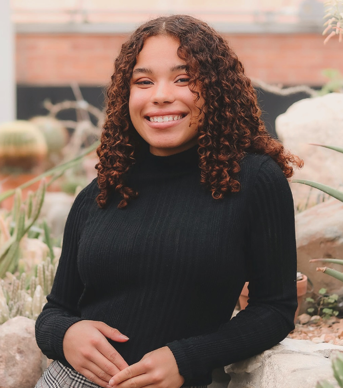 A smiling young woman with curly hair wearing a black top, standing in a botanical garden with cacti.