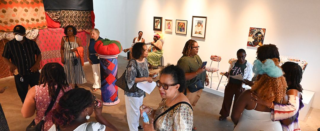 Attendees view textile art installations and framed paintings in a brightly lit art gallery.