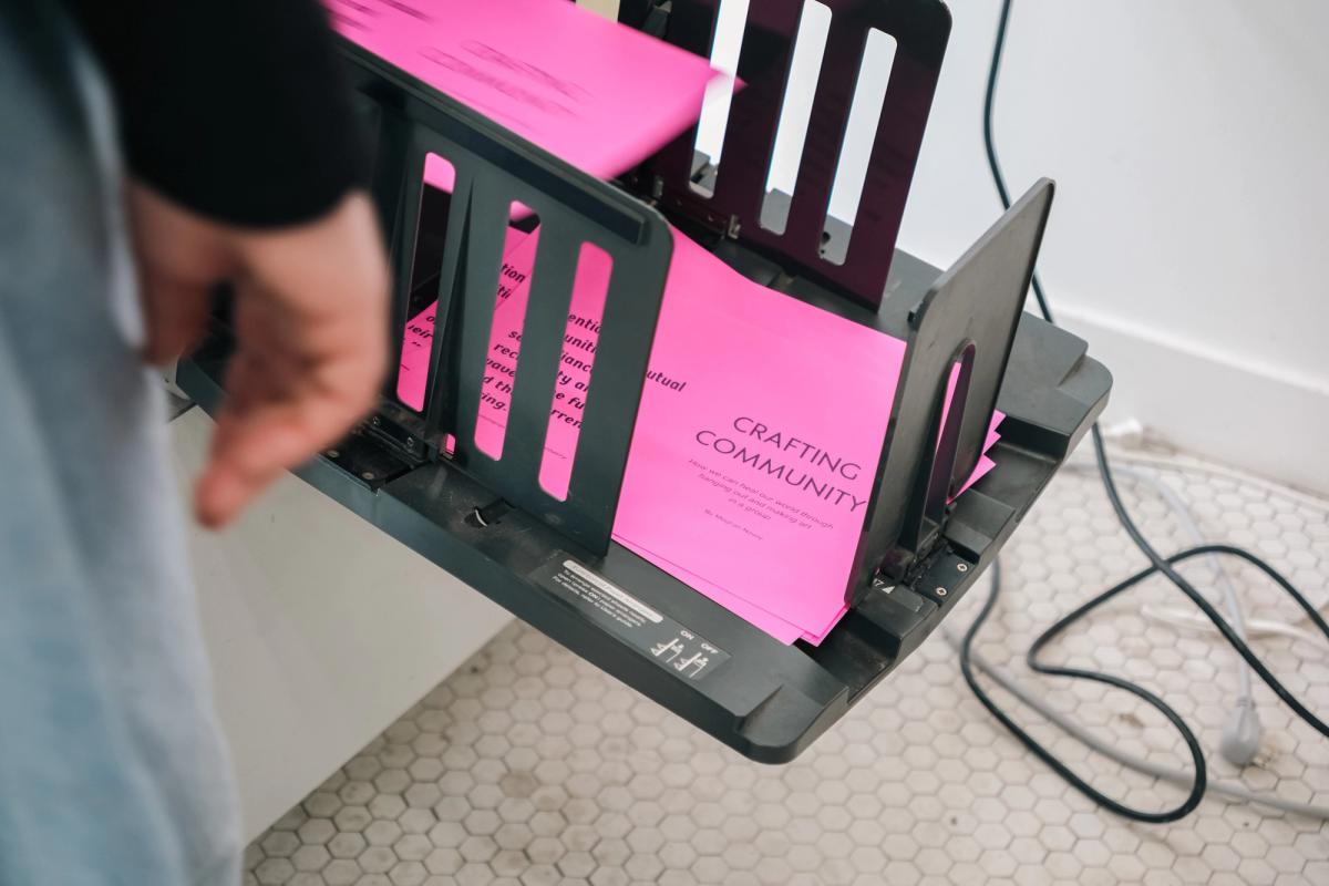 A person's hand next to a machine folding bright pink papers that read "CRAFTING COMMUNITY".