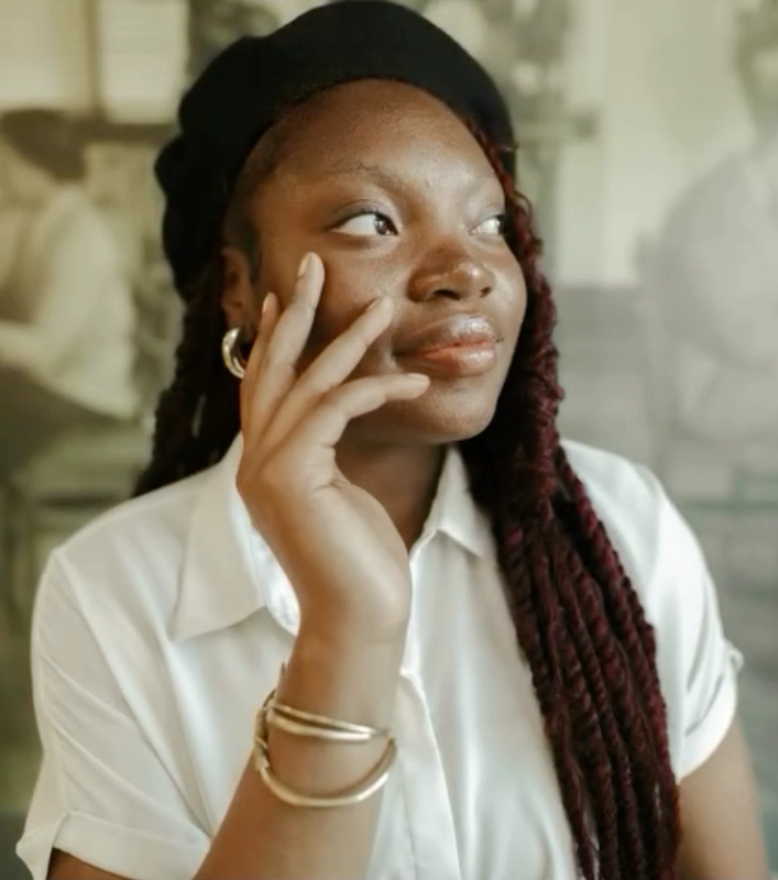 A young Black woman with long braids, wearing a black beret and white shirt, looks to the side with her hand on her face, against a blurry historical mural.