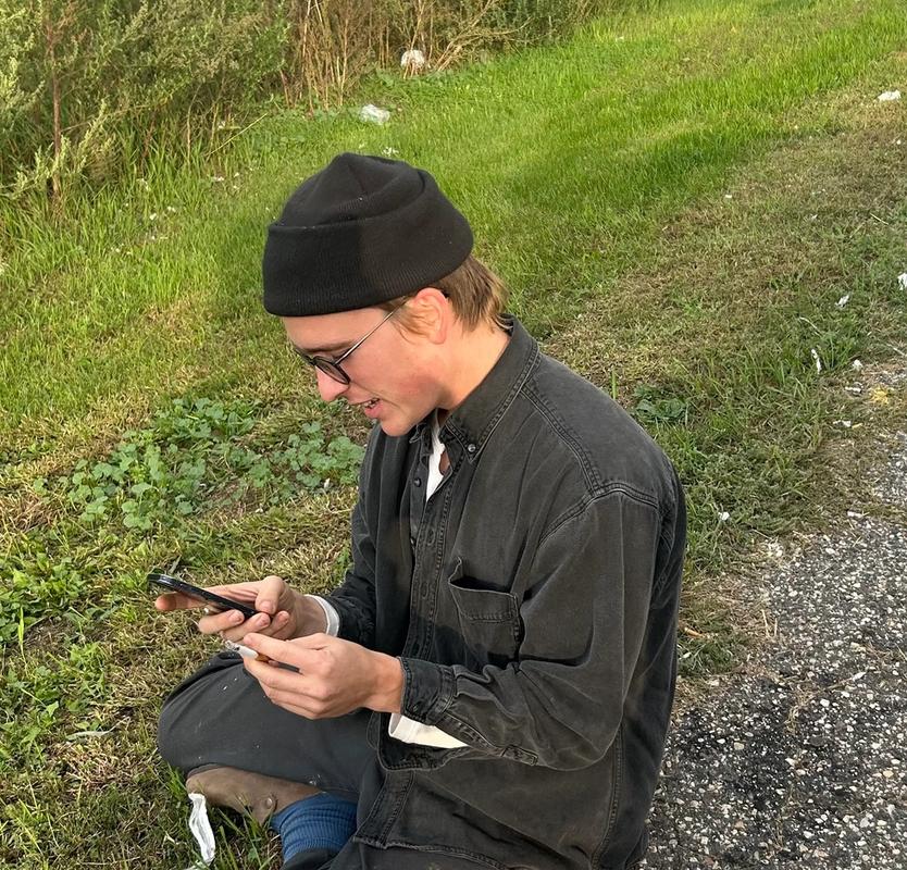 Young man in a black beanie and glasses crouches outdoors, looking at a phone while holding a cigarette pack.