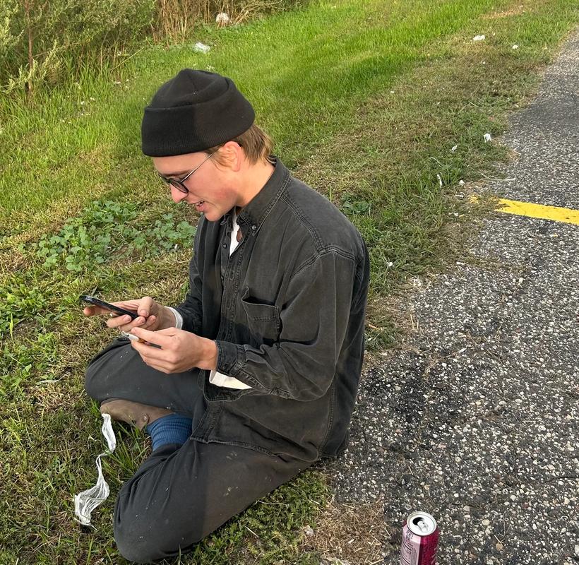 A young man in a black beanie and glasses sits cross-legged on the grass by an asphalt path, looking at his phone with a field in the background.
