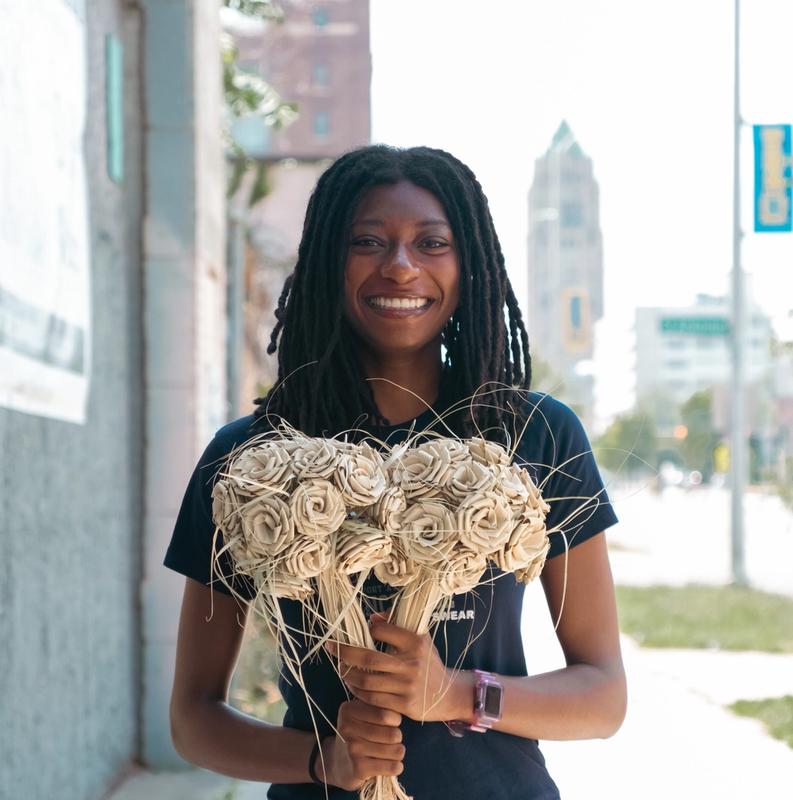 A smiling young Black woman with dreadlocks holds two bouquets of light-colored, dried craft flowers on a sunny city sidewalk.