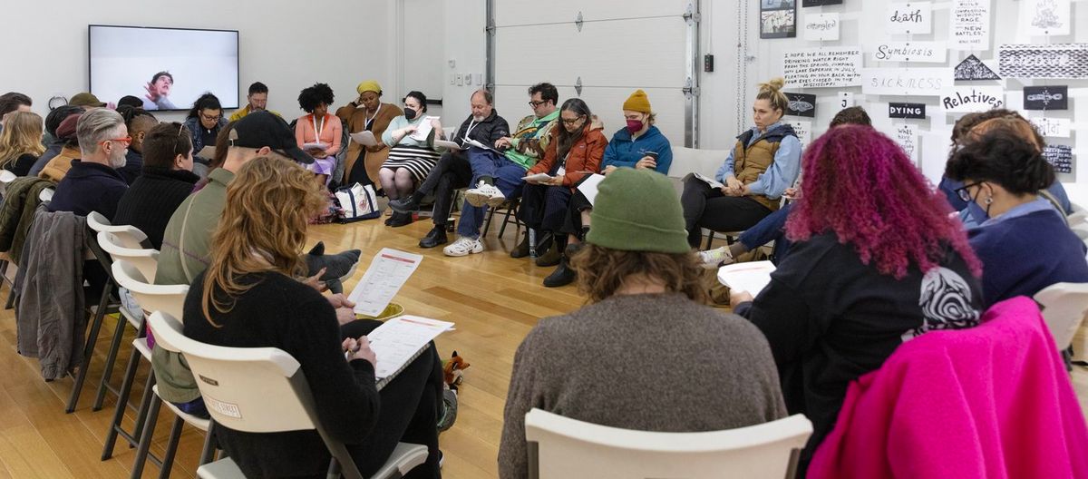 A diverse group of people meets in a circle in a bright, modern room, with a screen showing a person and a wall display featuring notes and a prominent yellow "ABOLISH" sign behind them.