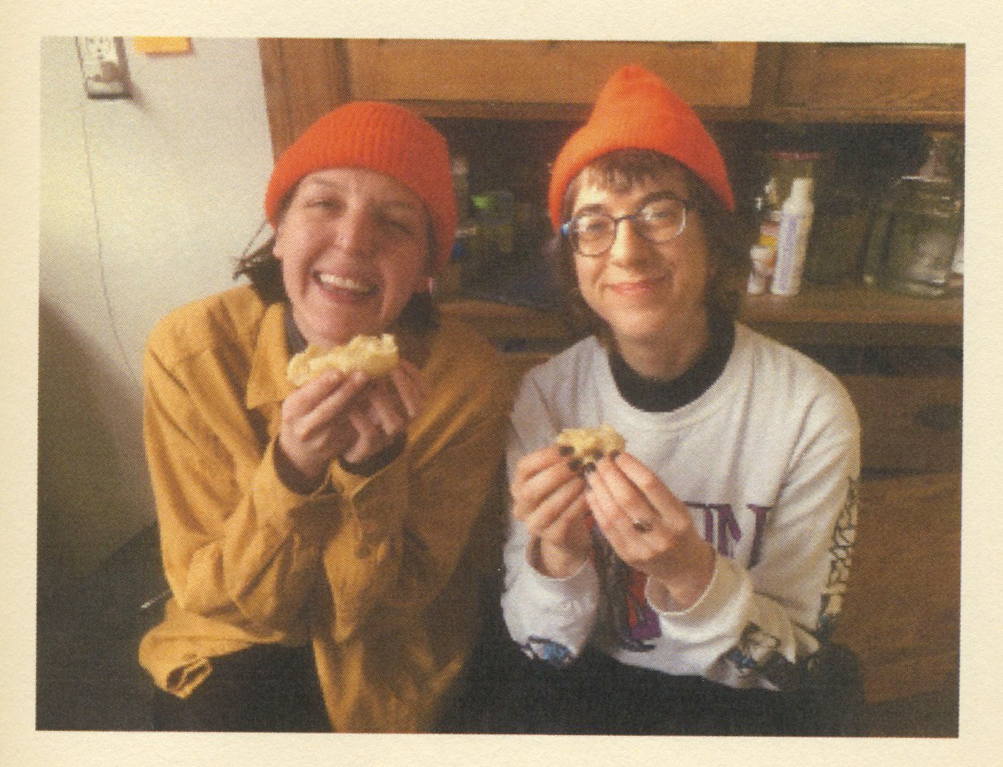 Two smiling people in orange beanies holding pastries.