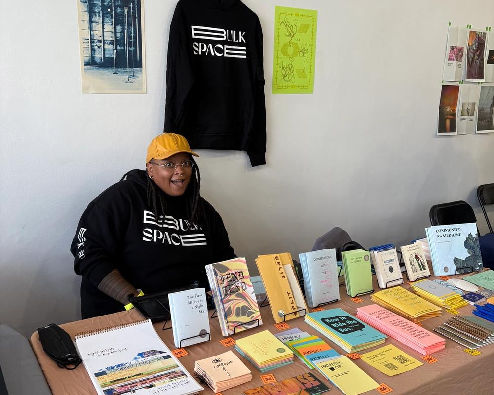A person in a yellow hat and black "BULK SPACE" hoodie sits smiling behind a table filled with various books and publications.