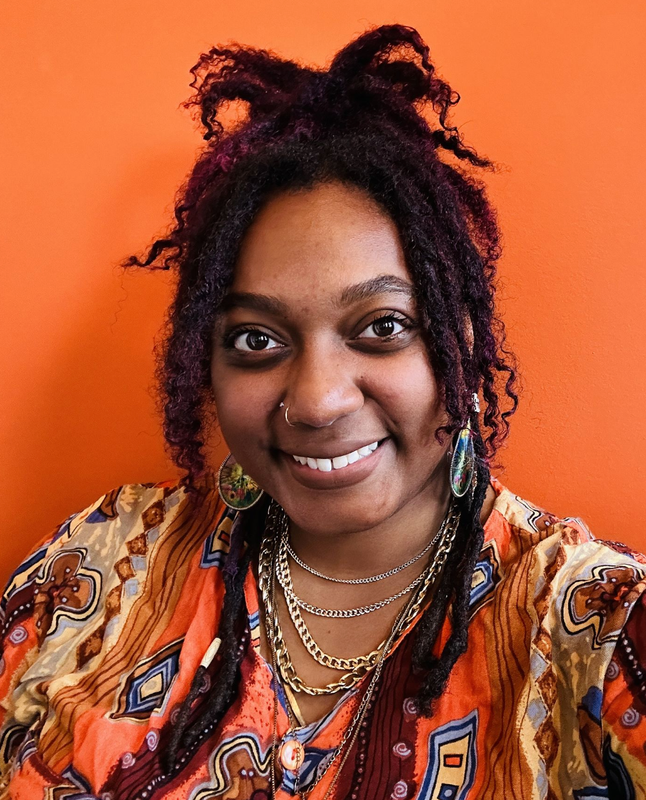 A smiling person with purple dreadlocks, layered necklaces, and a patterned shirt against an orange background.