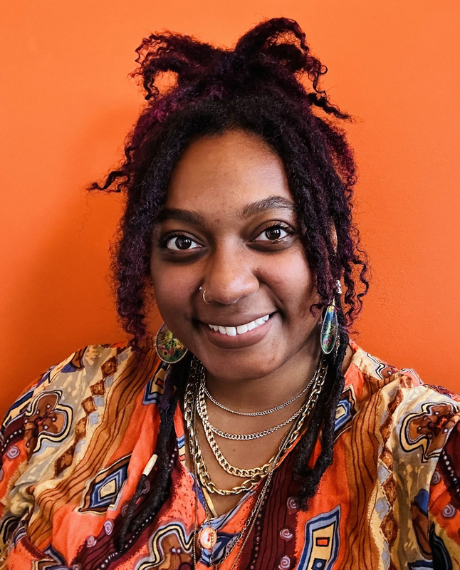 A smiling person with purple dreadlocks, layered necklaces, and a patterned shirt against an orange background.