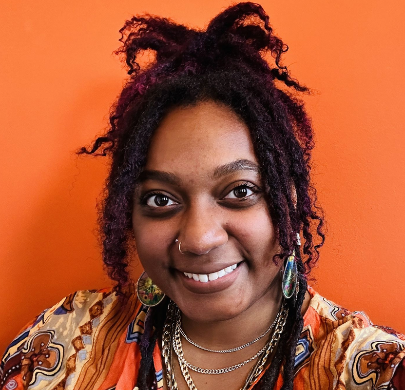 Smiling person with purple dreadlocks, layered necklaces, and a patterned shirt against an orange background.