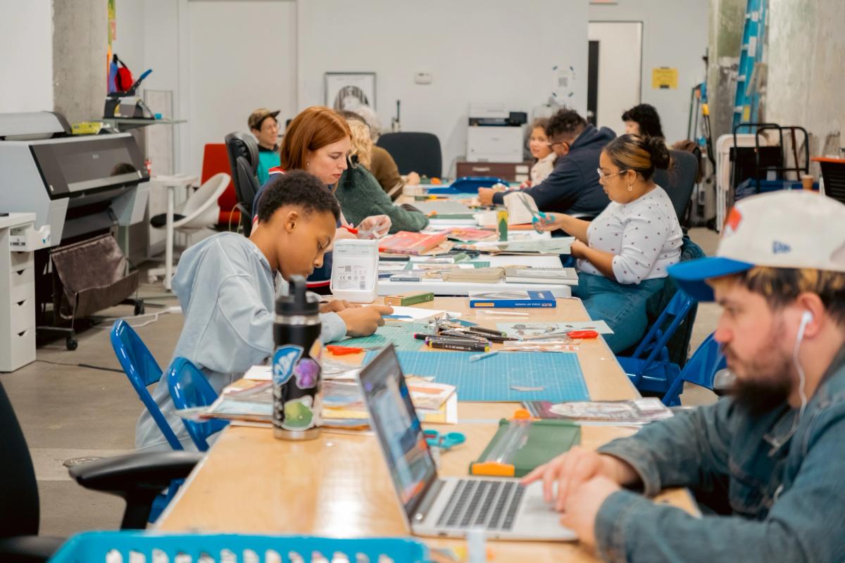 A diverse group of people works on crafts and laptops at long tables in a brightly lit workshop.