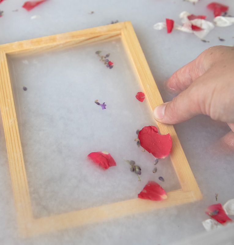 Hand holding a wooden papermaking screen in milky liquid with rose petals and lavender.